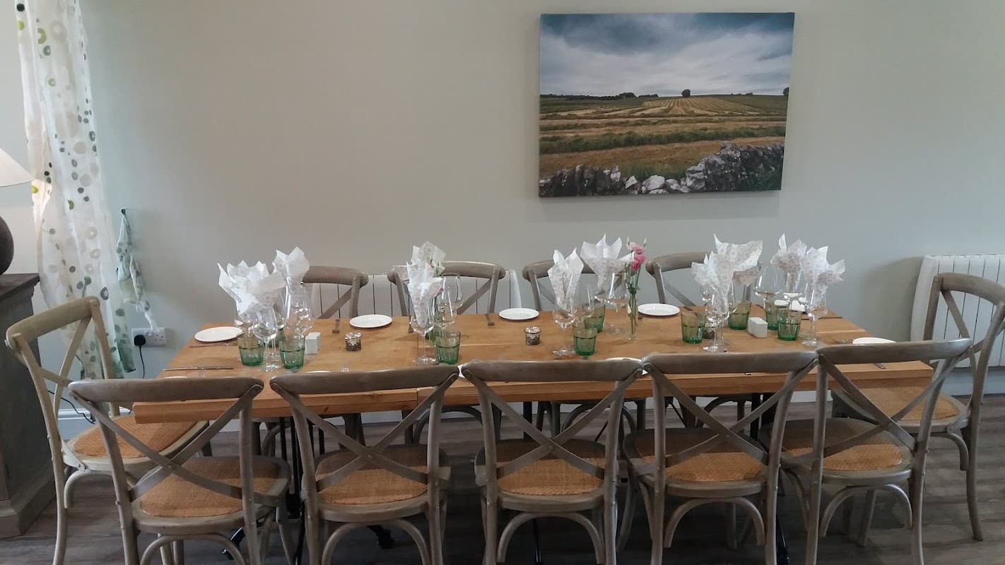 A long wooden dining table set with plates, glasses, and neatly folded napkins, against a scenic landscape painting.