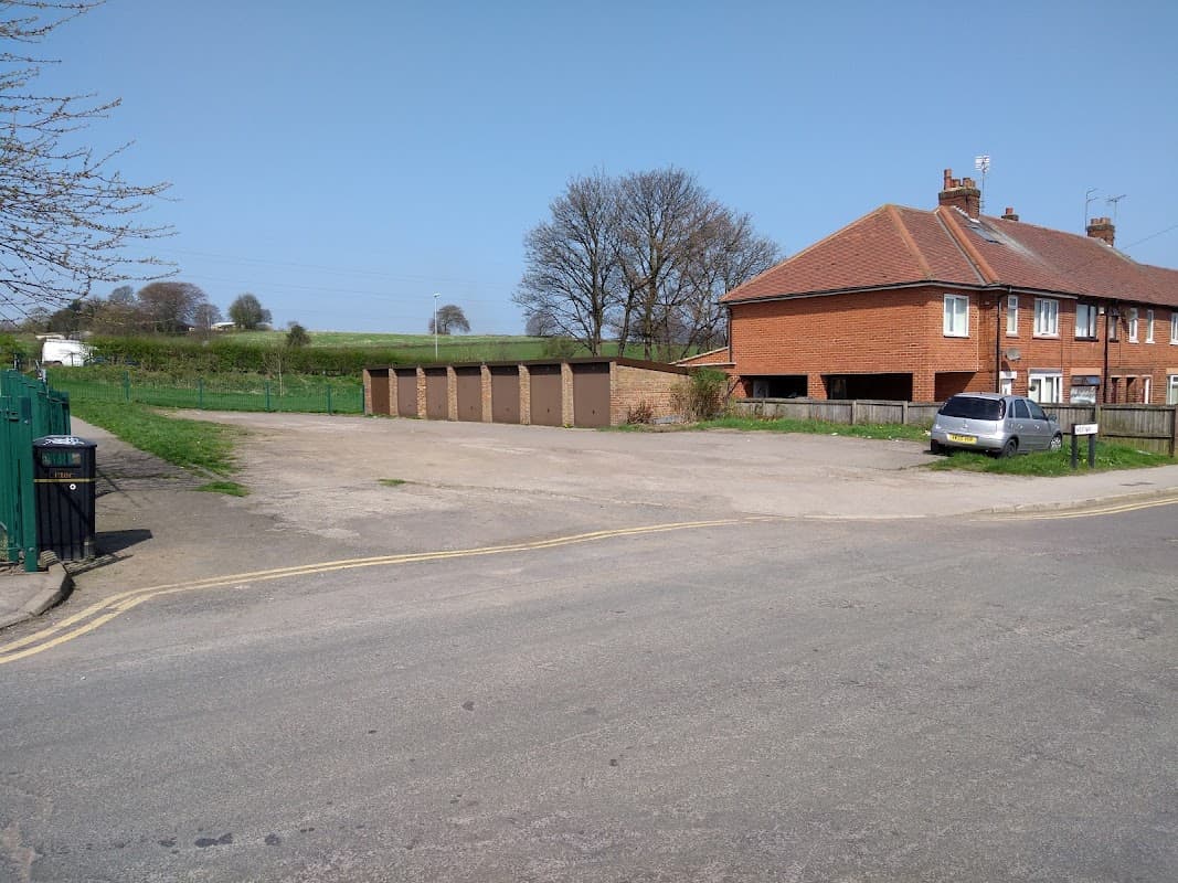 Pay & Display car park with garages, a residential building, and green fields in the background under a clear blue sky.