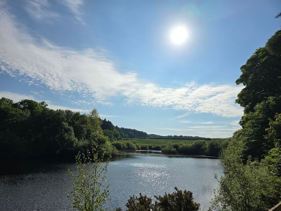 Bright sun above a serene reservoir surrounded by lush greenery and rolling hills under a partly cloudy sky.