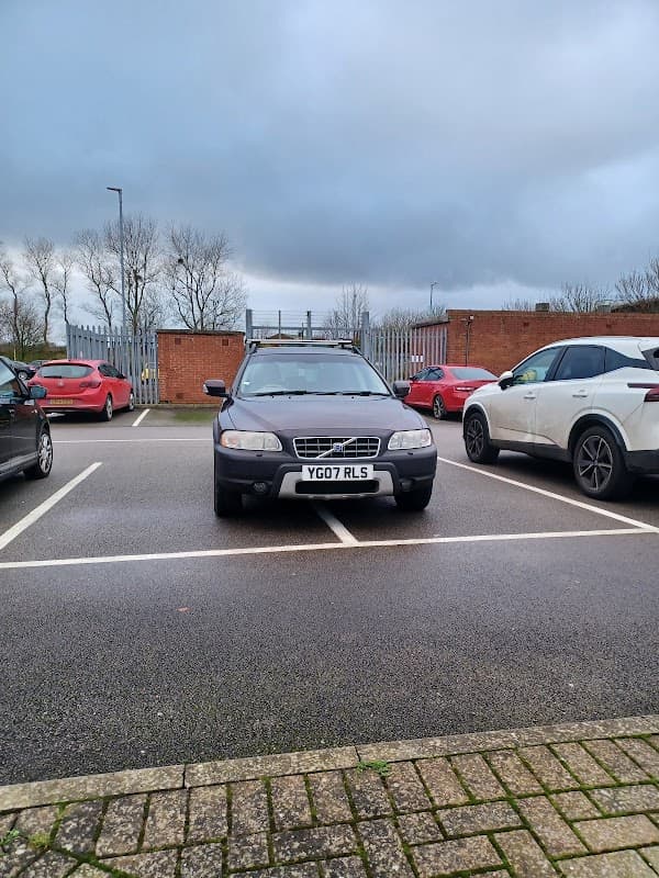 A black car parked across two spaces in a Pay & Display car park, with other cars and a cloudy sky in the background.