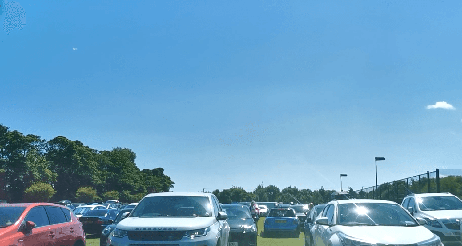 Busy car park filled with various vehicles under a clear blue sky, surrounded by greenery.