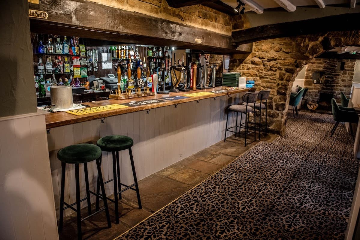 Bar area with wooden counter, stools, stone walls, and shelves lined with various drinks in The Queen's Head, Finghall.