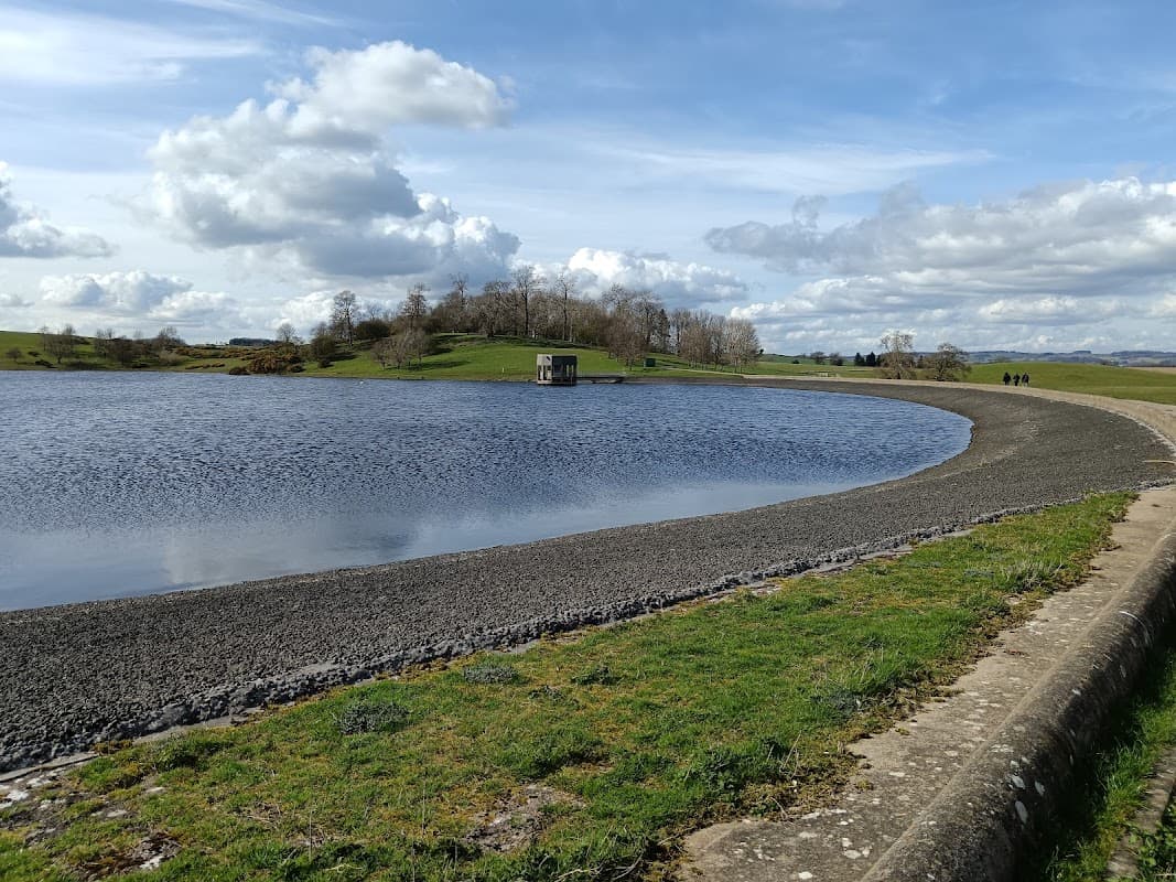 Car park near Thornton Steward Reservoir, with a calm water surface and grassy banks under a cloudy sky.