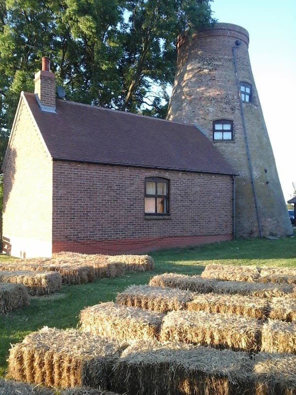 A brick building next to a tall, weathered mill tower, surrounded by straw bales and green grass.