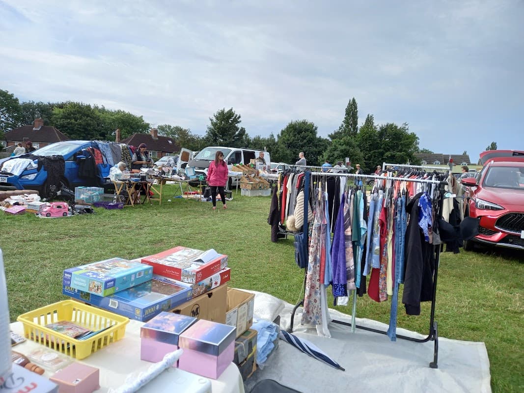 Vendors and shoppers browse various items at a busy car boot sale, with clothes hanging on a rack and parked cars nearby.