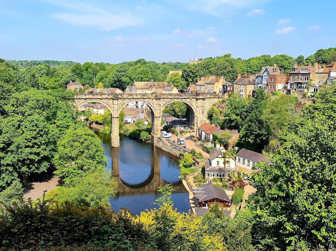 Scenic view of Knaresborough with a river, lush greenery, and a historic stone viaduct in the background.