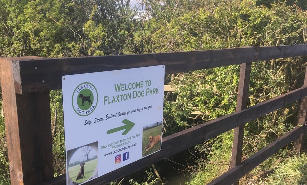 Welcome sign for Flaxton Dog Park on a wooden fence, surrounded by greenery and a view of the park beyond.