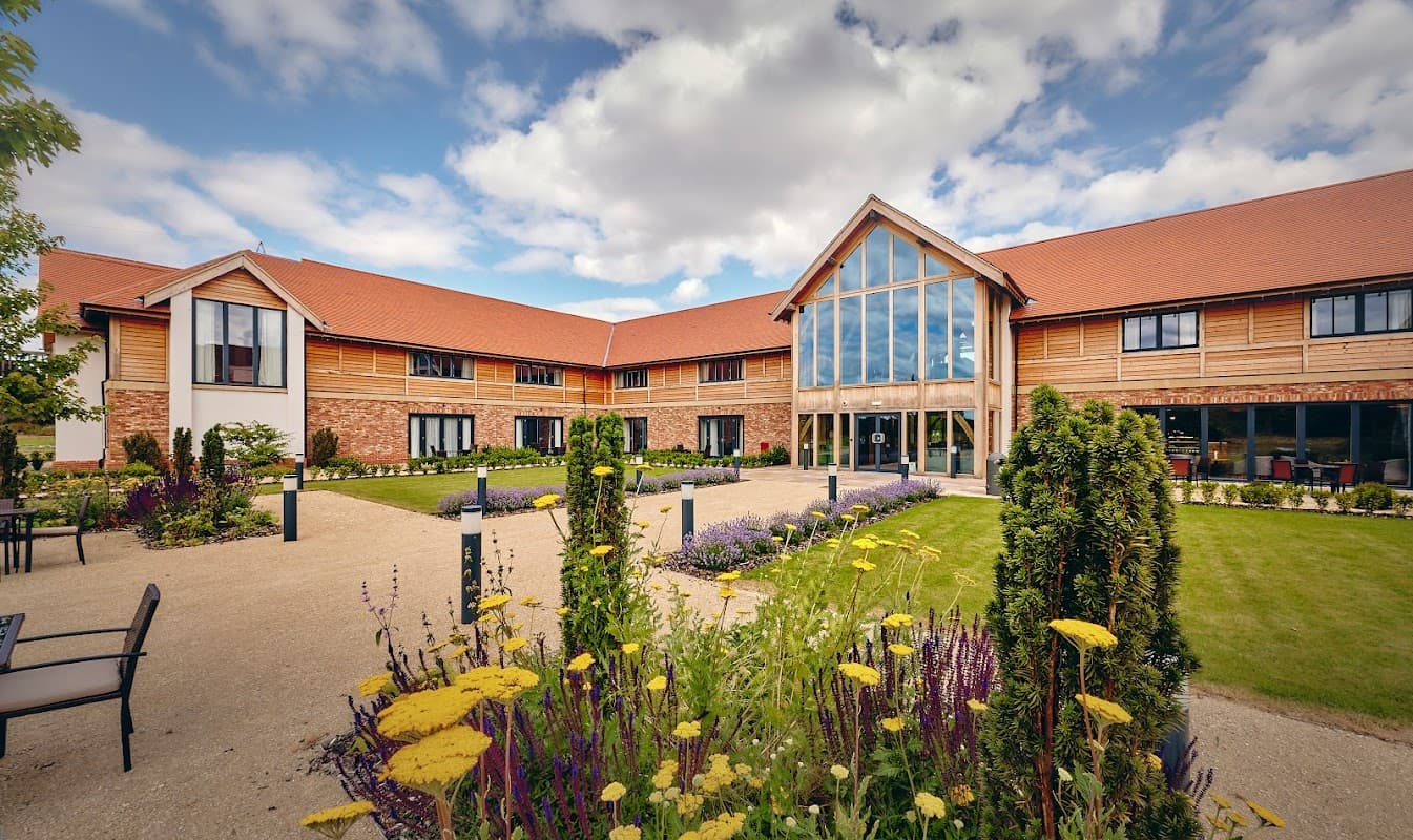 Modern restaurant with large windows, surrounded by landscaped gardens and colorful flowers under a partly cloudy sky.