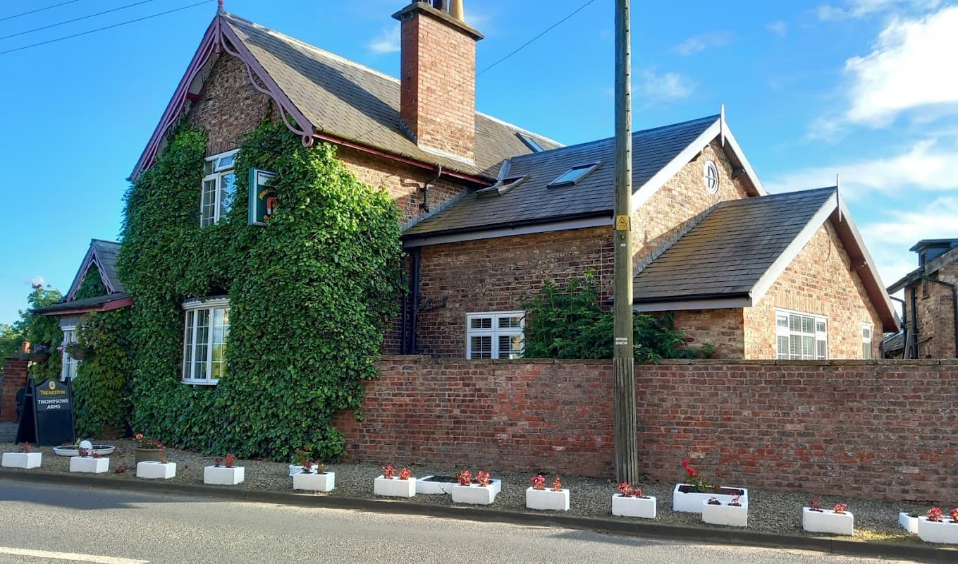 Brick building with ivy-covered walls, white window frames, and a flower bed along the front, set against a blue sky.