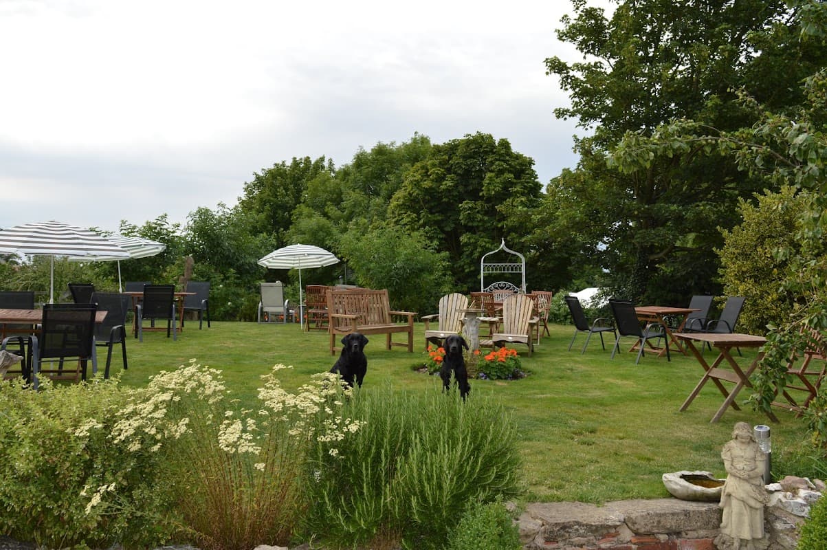 Outdoor seating area with wooden tables, chairs, and umbrellas surrounded by greenery and flowering plants.