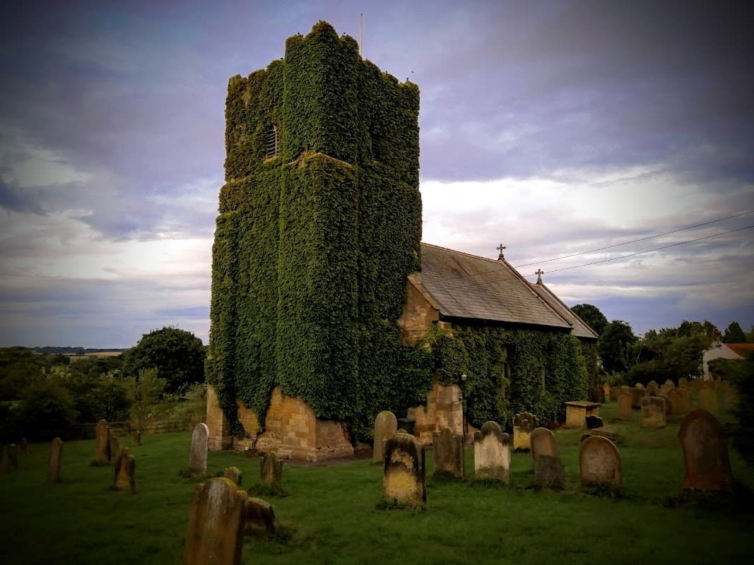 St. John The Evangelist Church covered in ivy, surrounded by gravestones and lush greenery under a cloudy sky.