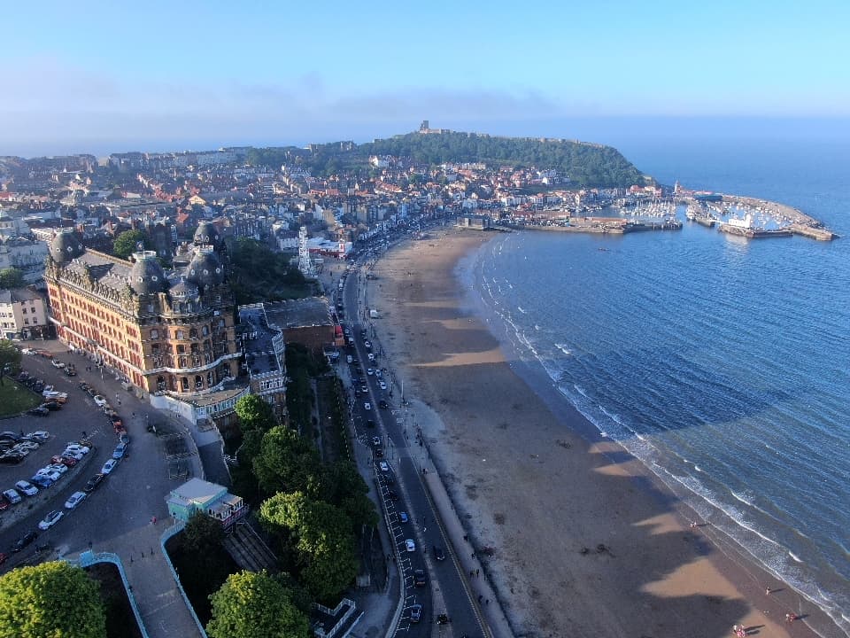 Aerial view of a coastal town with sandy beach, harbor, and buildings, under a clear blue sky.