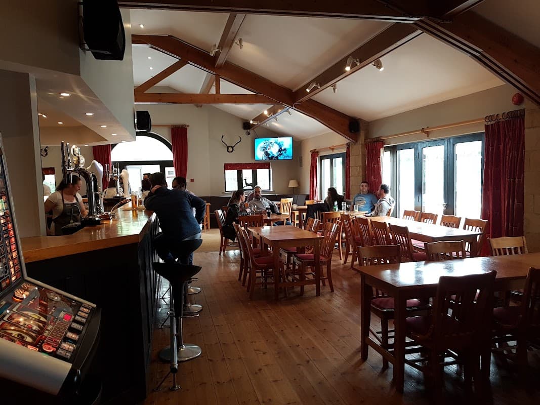 Warm pub interior with wooden beams, tables, and patrons enjoying drinks. A TV displays a sports game in the background.