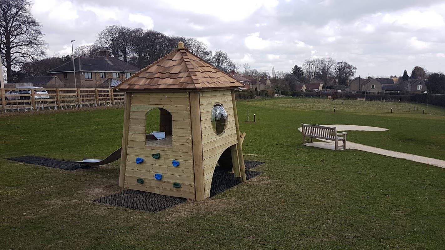 Wooden playhouse with a shingled roof, climbing rocks, and a slide in a grassy area with benches and trees.