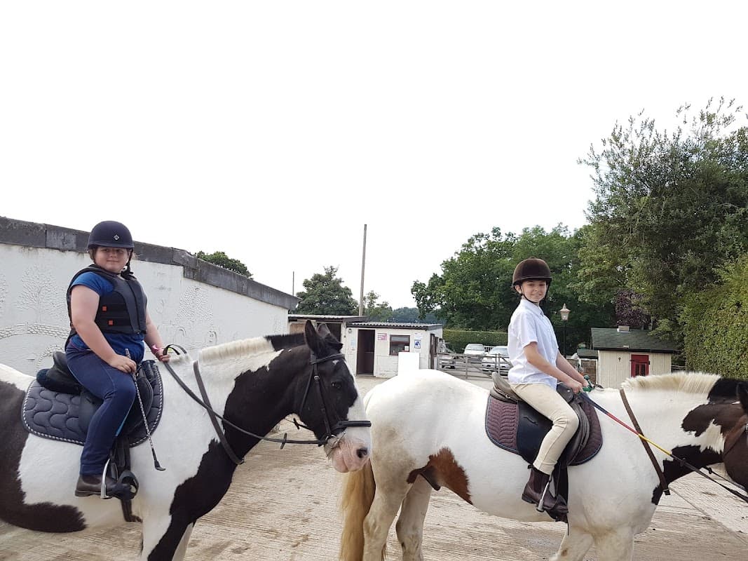 Two riders on horses at Follifoot Park Riding Centre, surrounded by trees and a stable building in the background.