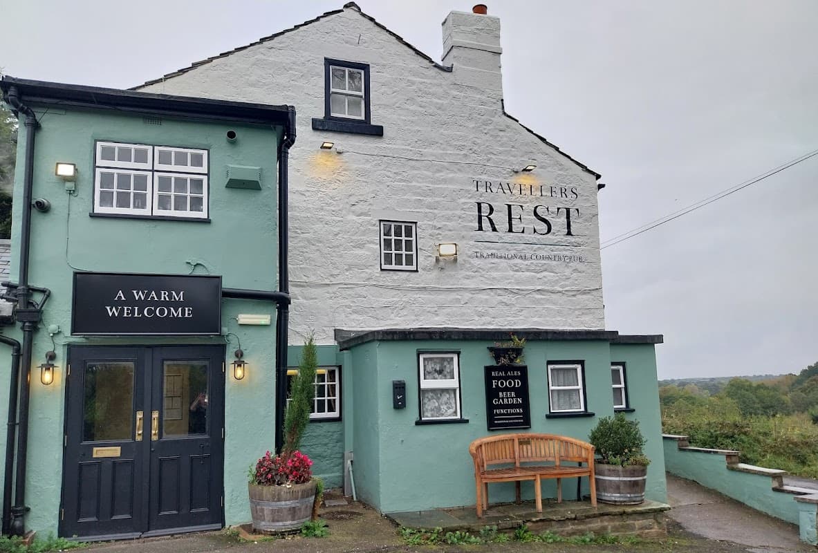 The Travellers Rest pub features a cozy exterior with a welcoming sign, flower pots, and a bench outside.
