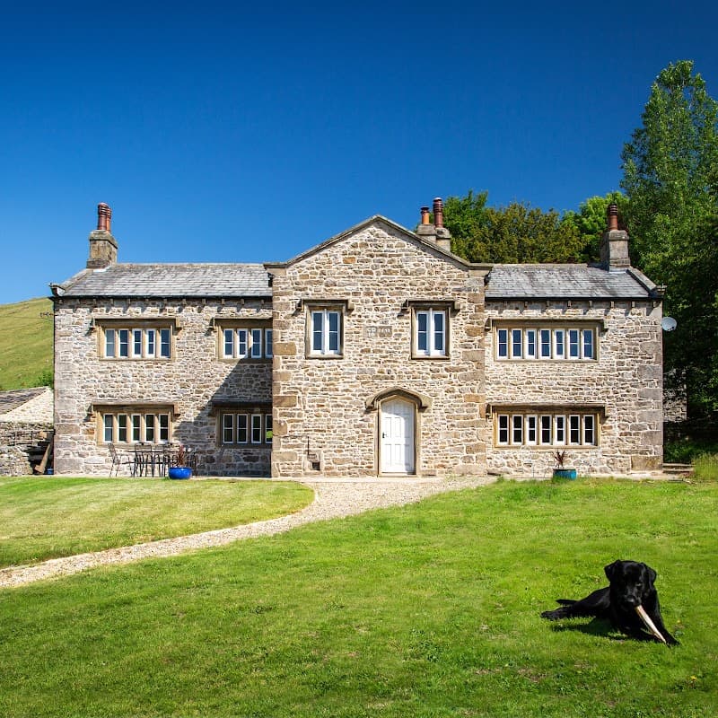 Stone building with large windows, set in a green landscape, featuring a black dog resting on the grass.