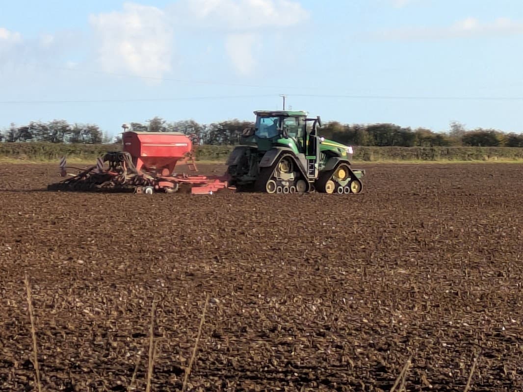 A green tractor with a red seed drill is working in a plowed field under a clear blue sky.