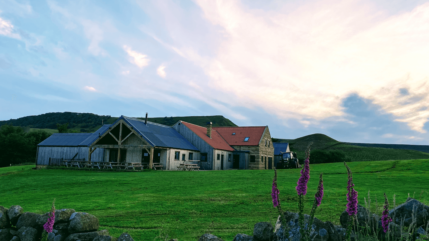 Yorkshire Cycle Hub cafe with rustic buildings, green fields, and a backdrop of rolling hills under a colorful sky.
