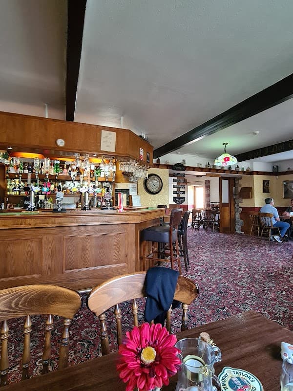 Cozy pub interior featuring a wooden bar, colorful drinks, floral decor, and patrons seated at tables on patterned carpet.