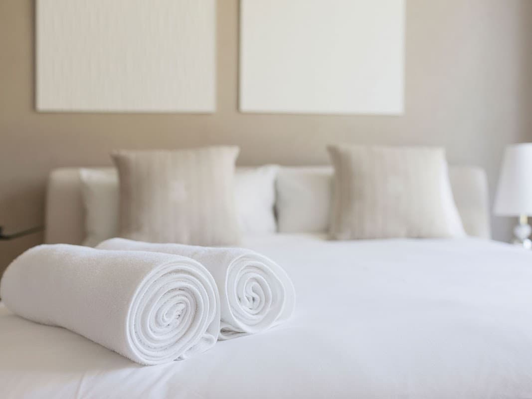 Neatly rolled white towels on a crisp bed with soft pillows against a neutral wall.