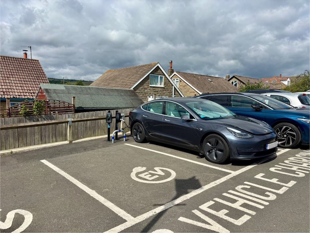 Connected Kerb Charging Station with a parked electric vehicle in a parking lot, surrounded by houses and cloudy skies.
