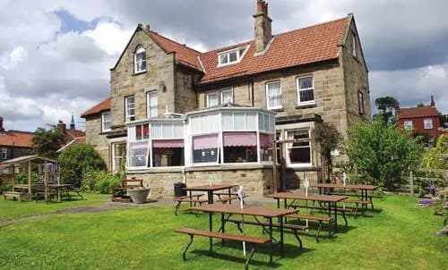 Fylingdales Inn features a stone building with a red-tiled roof, outdoor seating, and a grassy area under a cloudy sky.