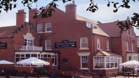 Red-brick building with multiple windows, outdoor seating, and a sign for Grosvenor Hotel in a quaint village setting.