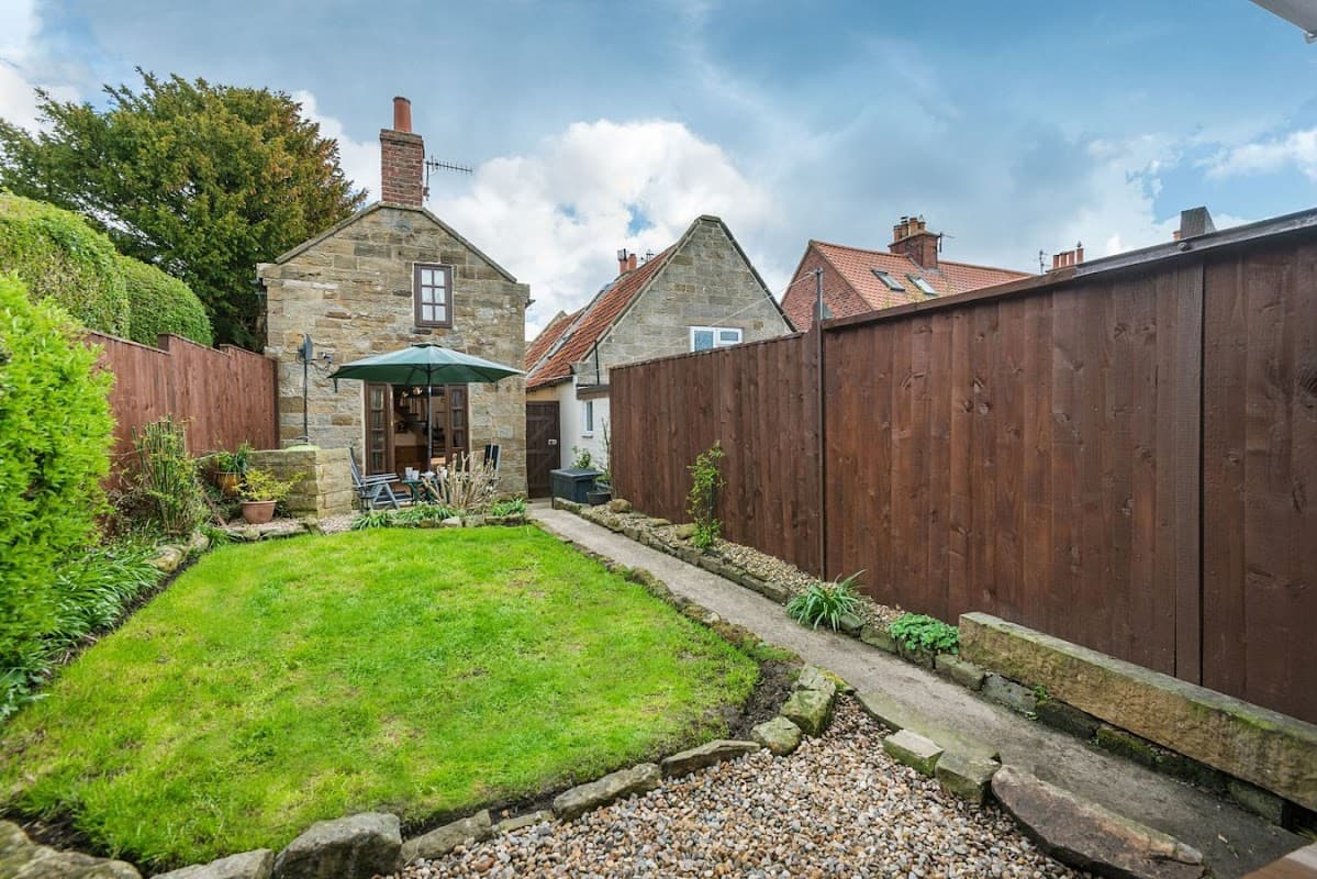 Charming stone cottage with a green lawn, patio furniture, and wooden fencing in a peaceful garden setting.