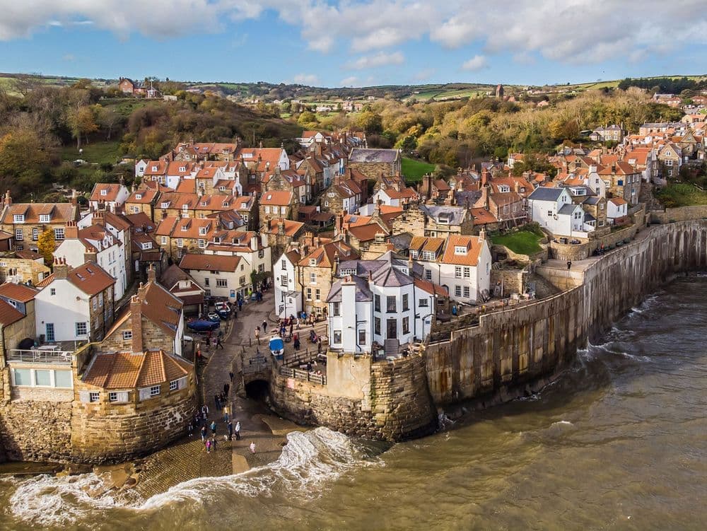 Charming coastal village with red-roofed cottages, rocky shoreline, and a scenic backdrop of hills and blue sky.