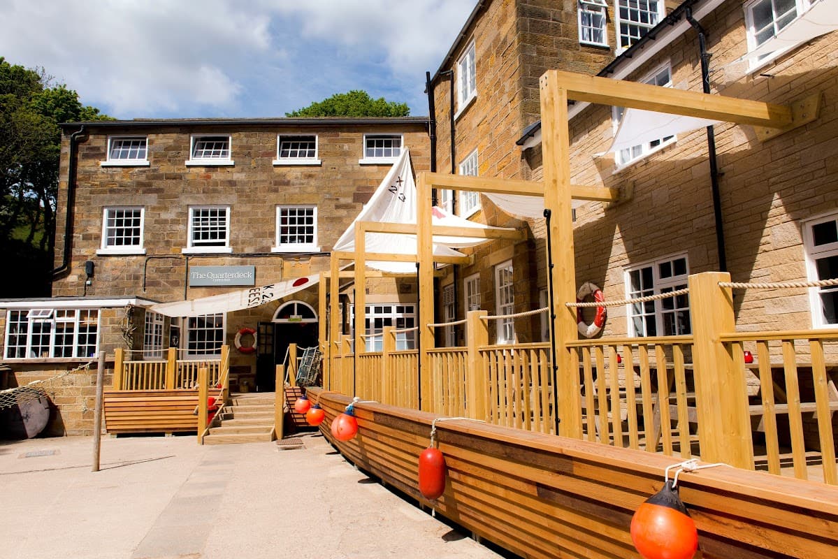 Stone building with wooden deck, lifebuoys, and white awnings under a blue sky in a coastal setting.