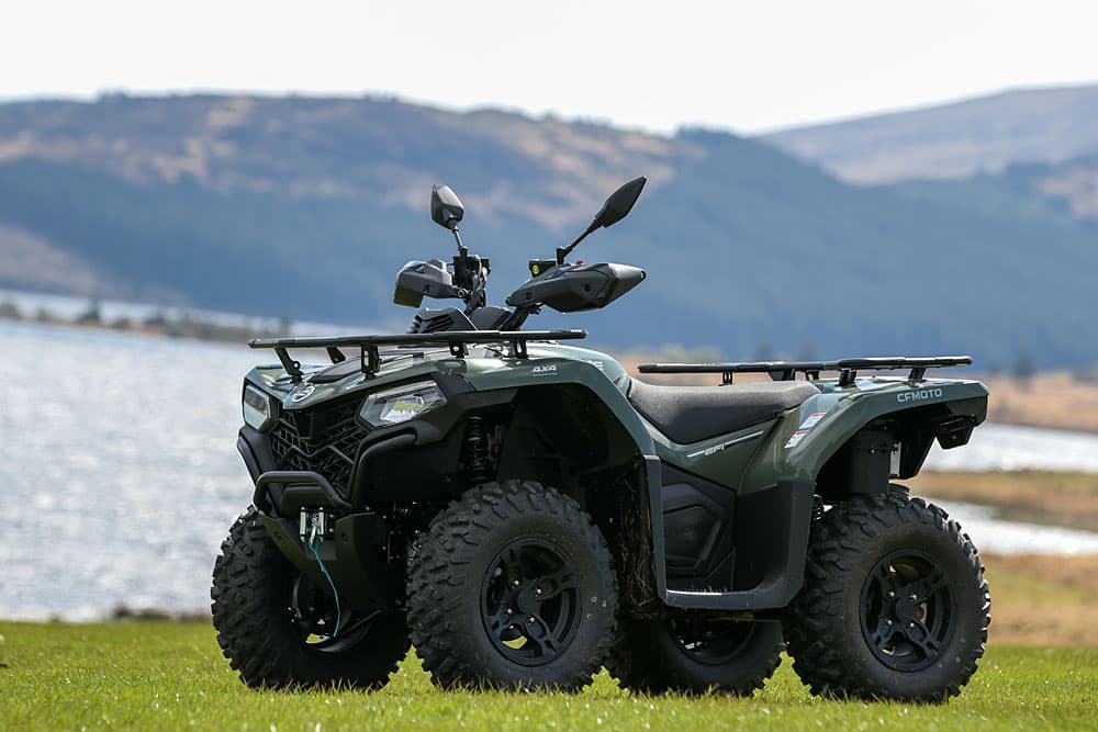 A green utility vehicle parked on grass near a lake, with hills and trees in the background.