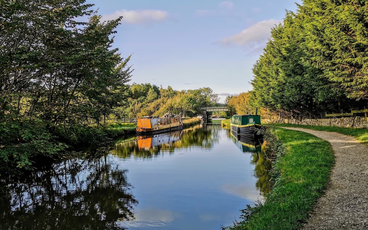 Calm canal scene with moored boats, lush greenery, and a path along the water under a clear blue sky.