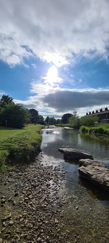 Sunlight reflects on a tranquil river, surrounded by greenery and stone, with buildings visible in the background.