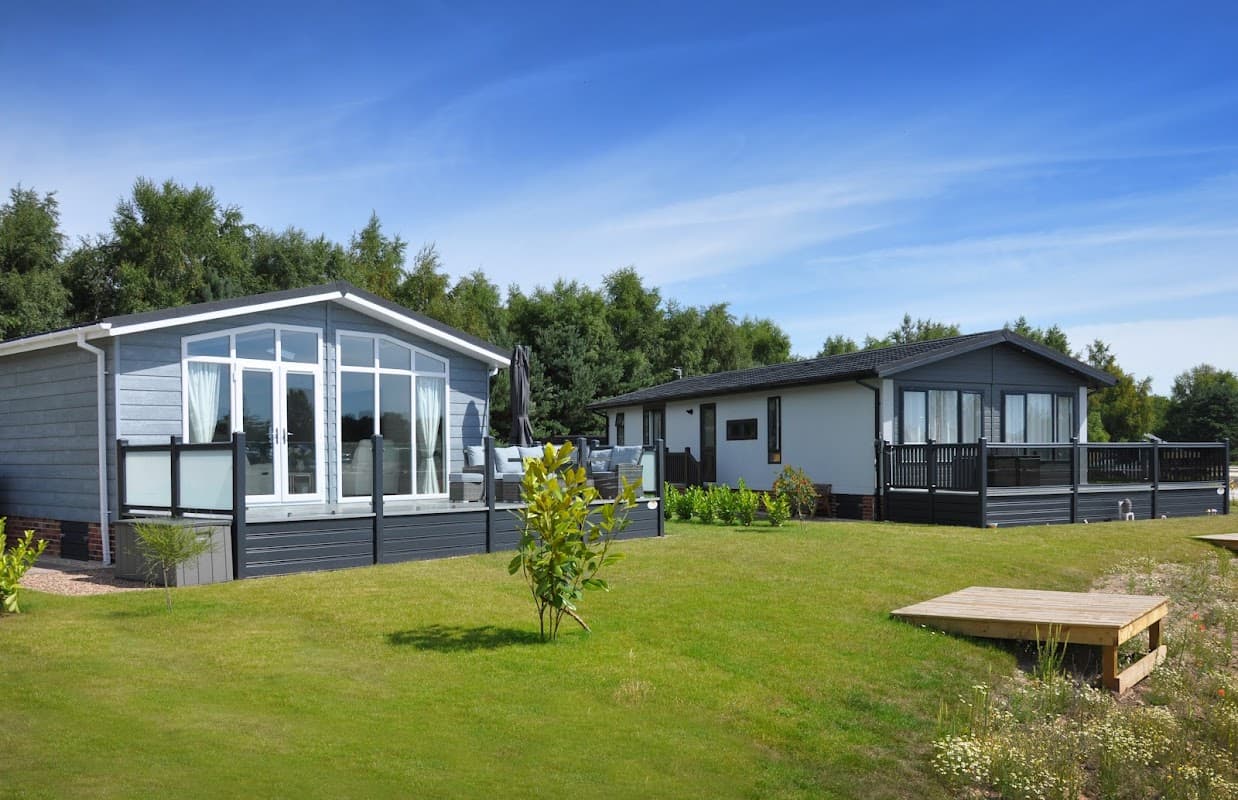 Two modern lodges with large windows on a grassy area, surrounded by trees under a clear blue sky.