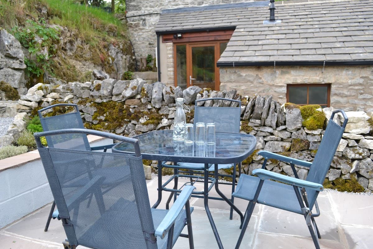 Outdoor seating area with a glass table and chairs, surrounded by stone walls and greenery at Ashes Farm.