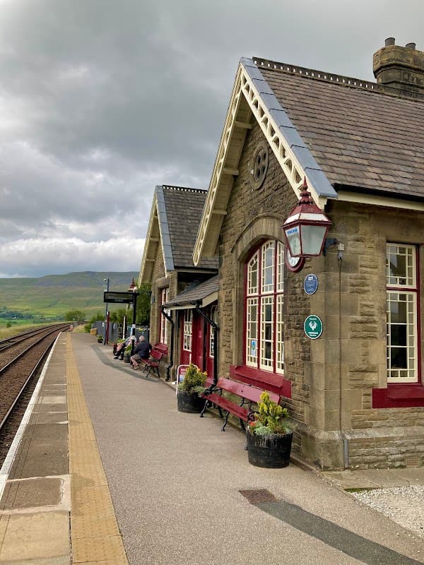Ribblehead Station features a stone building with red accents, benches, and scenic views of hills under a cloudy sky.
