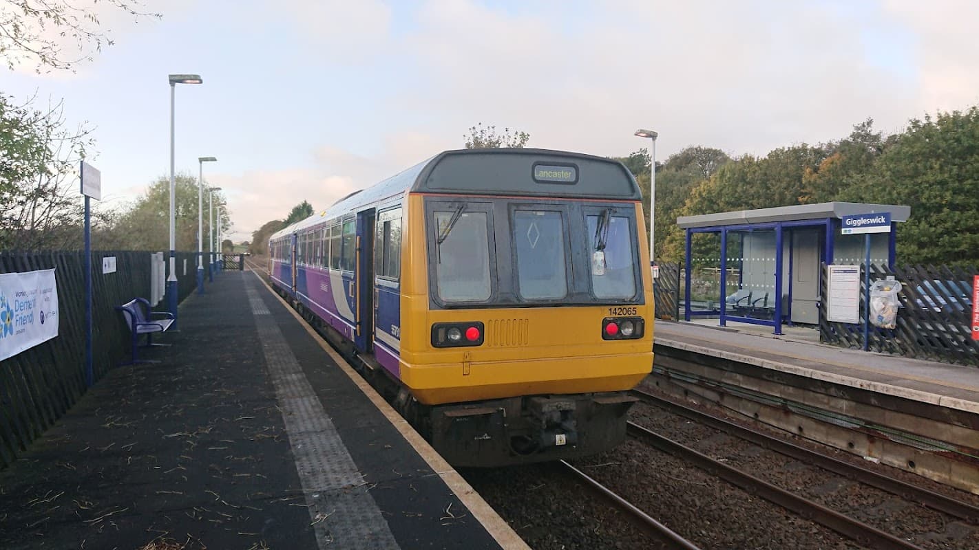 A train at Giggleswick station, with platform seating and signage amidst a green landscape.