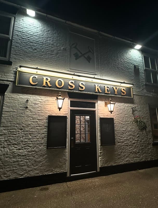 Illuminated sign reading "CROSS KEYS" on a brick pub exterior with two wall-mounted lanterns at night.