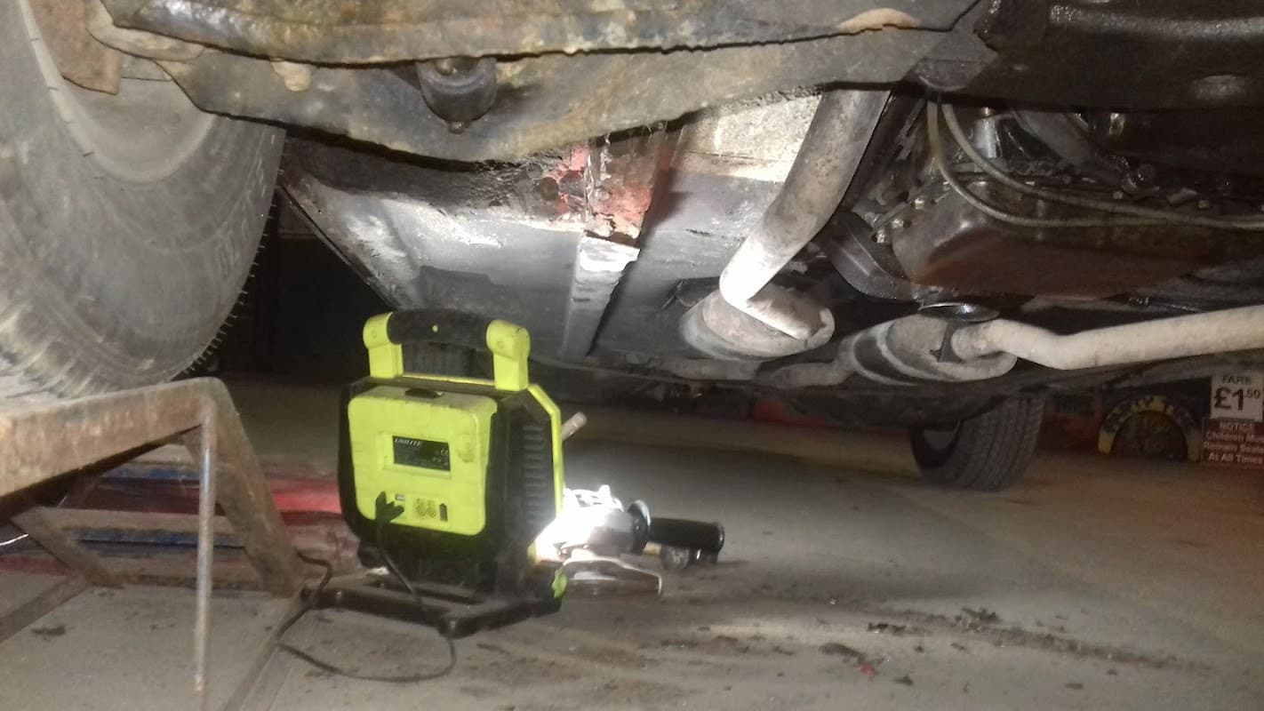 Underneath a car, showing a partially visible undercarriage and tools on the ground, with a bright work light nearby.
