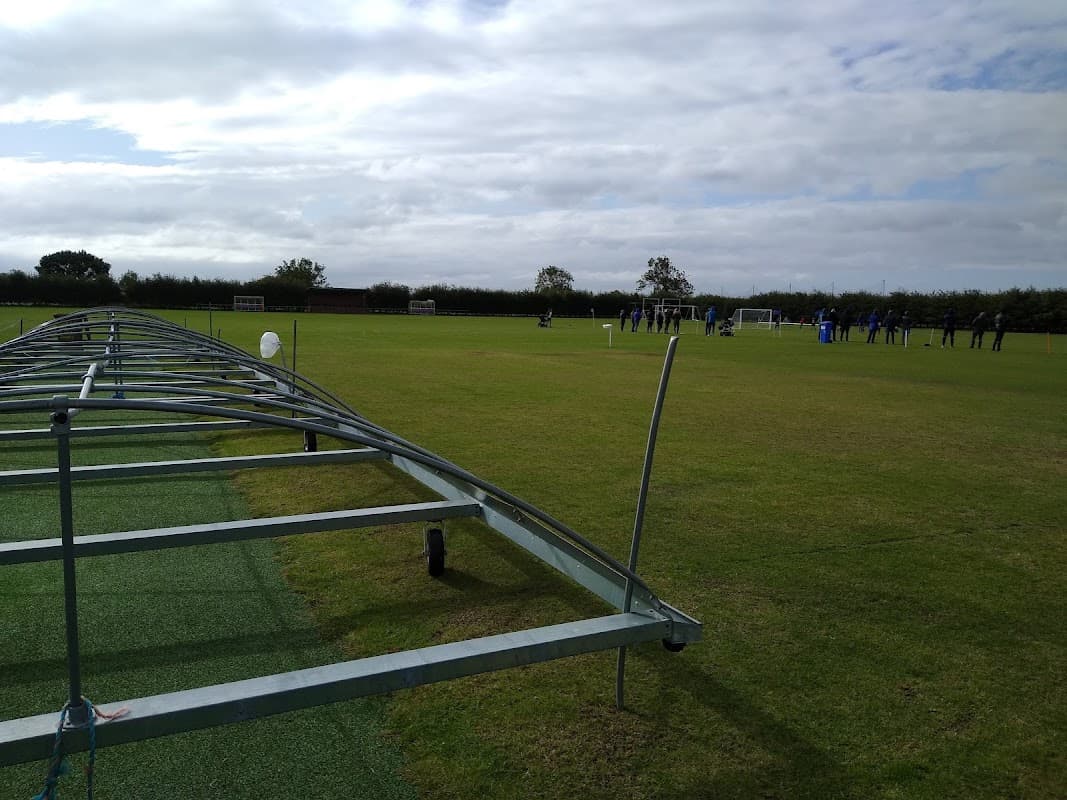Sports field with green grass, goalposts, and players in training under a cloudy sky.
