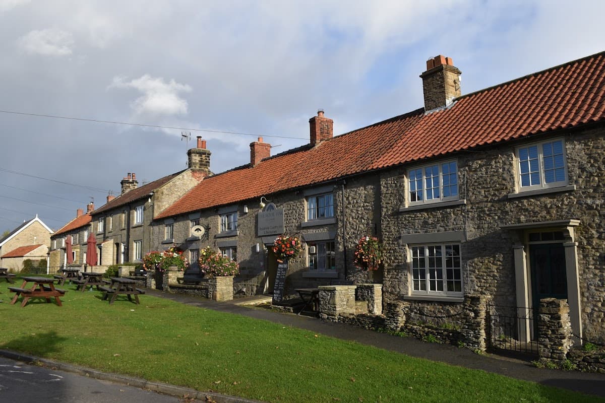 Charming stone building with a red-tiled roof, flower baskets, and picnic tables outside in a quaint village setting.