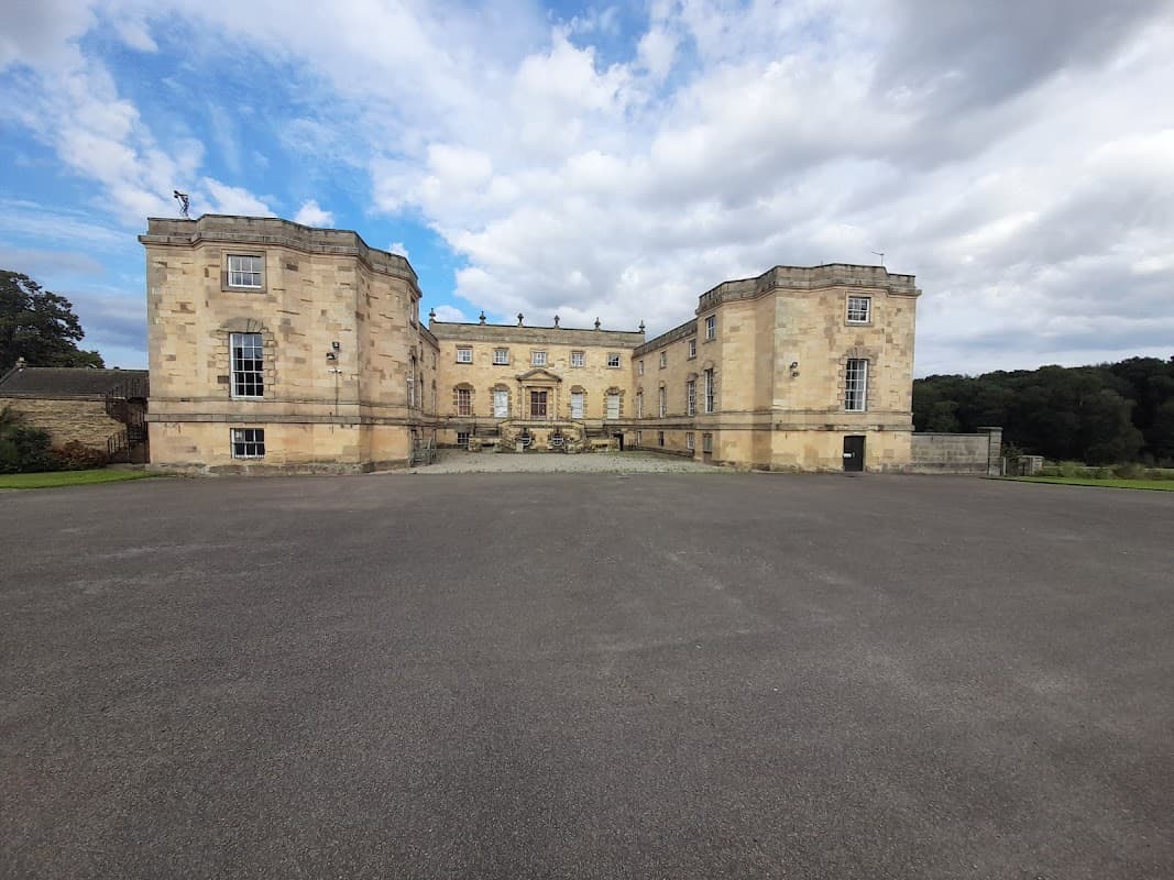 Gilling Castle with stone facade and large central entrance, surrounded by a paved area and lush greenery in the background.