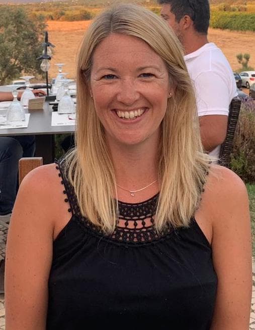 Smiling woman with long blonde hair, wearing a black top, seated outdoors with a table set in the background.