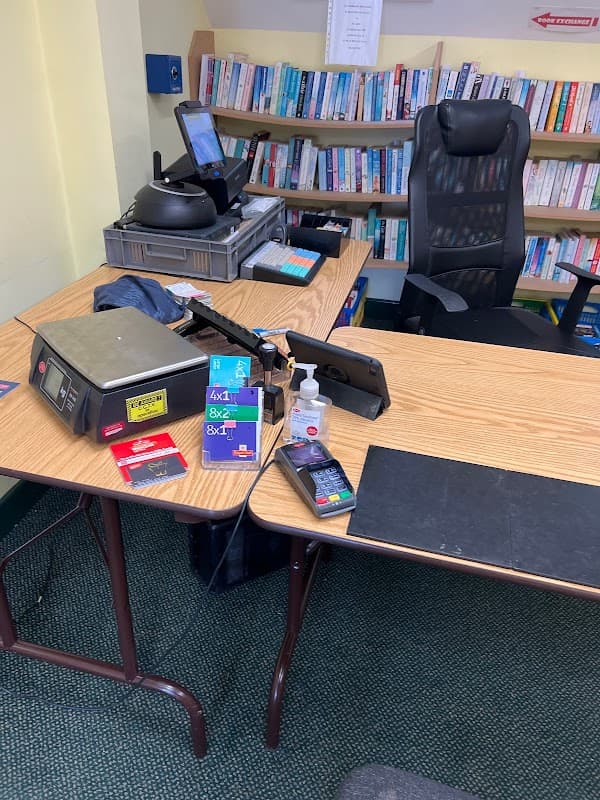 A small post office counter with a cash register, payment terminal, hand sanitizer, and a chair in front of a bookshelf.