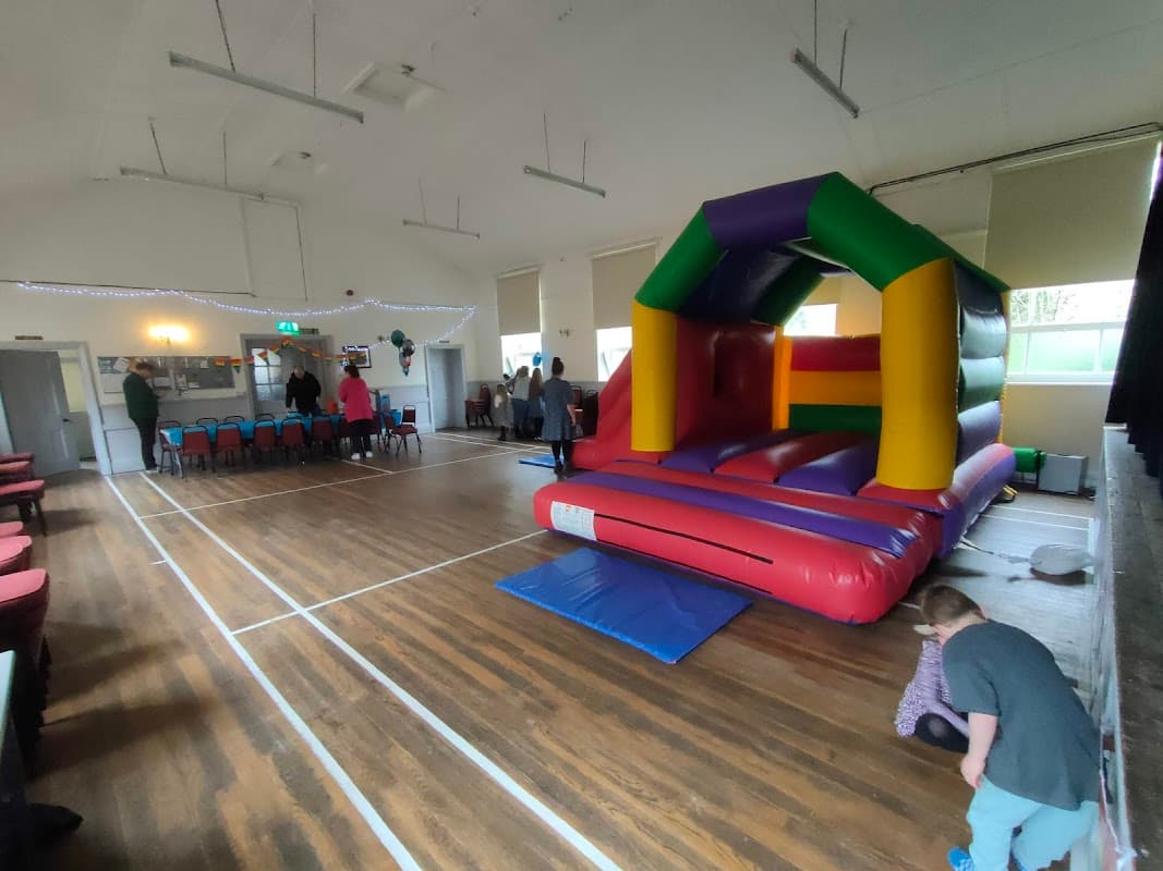 Colorful bouncy castle in a hall with wooden floors and chairs arranged in the background, people interacting nearby.