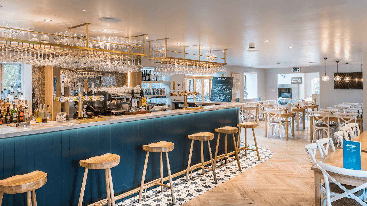 Stylish bar with wooden stools, glassware, and a bright dining area featuring wooden tables in a modern setting.