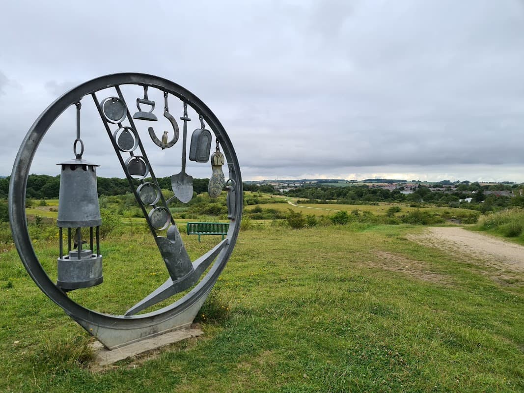 Metal sculpture featuring various tools, with a grassy landscape and cloudy sky in the background.