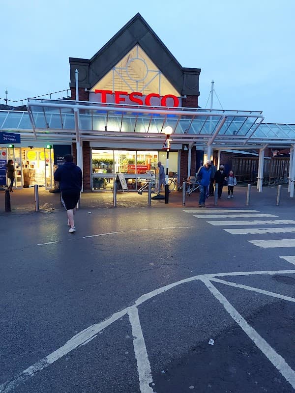 Tesco Superstore entrance with shoppers, illuminated sign, and a cloudy evening sky in Goole, Yorkshire.