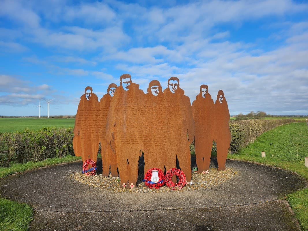 Silhouetted metal figures stand in a circle, surrounded by wreaths, against a blue sky and green fields.